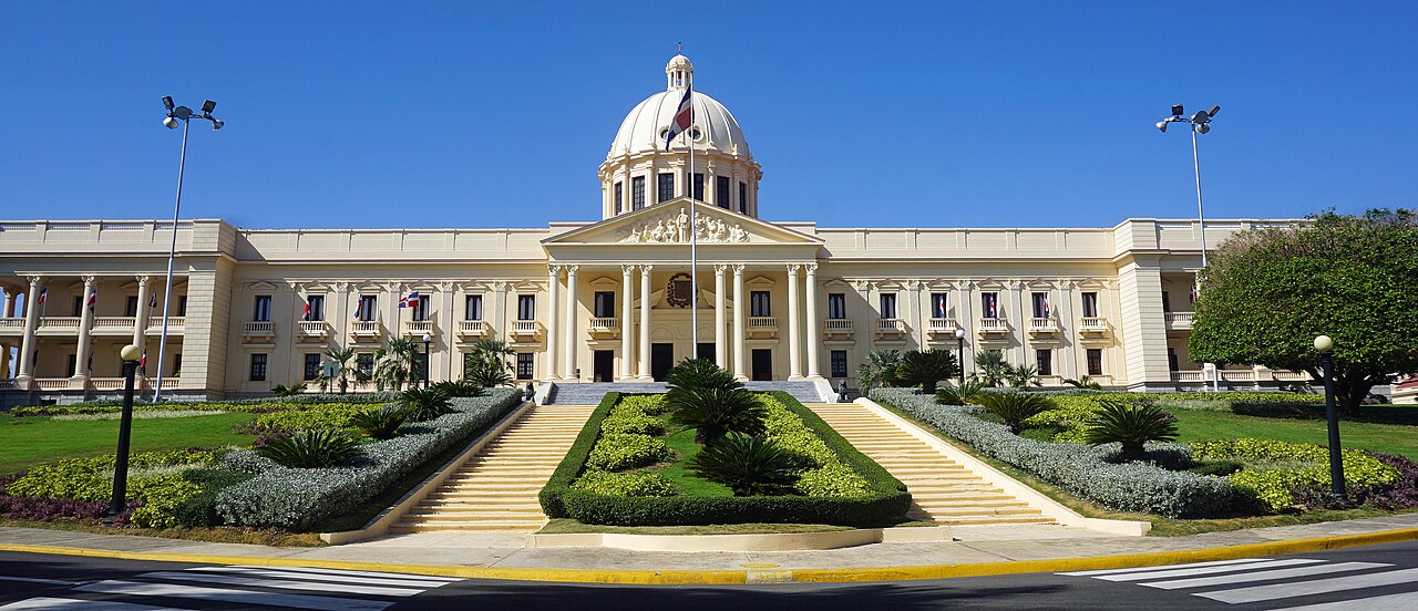 A cream-coloured domed palace with a columned entrance at the top of a broad staircase flanked by clipped gardens.
