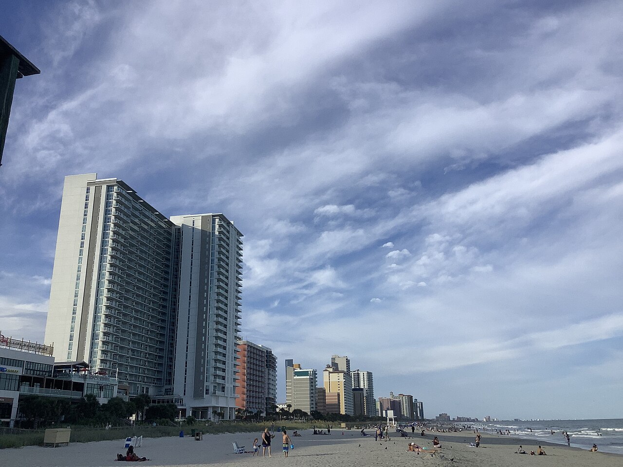 A line of high-rise hotel towers running along a beach into the distance under a sky of streaked cloud.