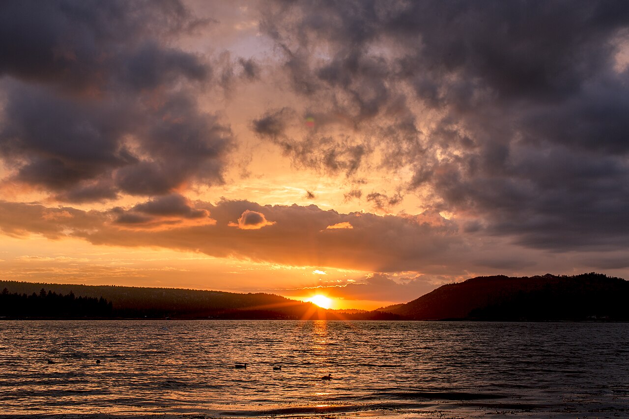 An orange sun setting behind dark hills across a lake under heavy clouds.