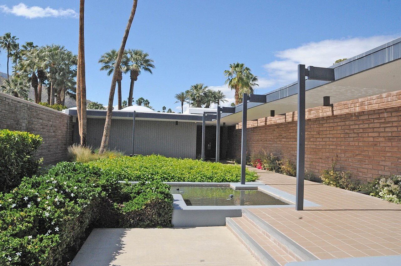 A low modernist house with a flat roof and brick wall behind a clipped garden, a reflecting pool and tall palms.