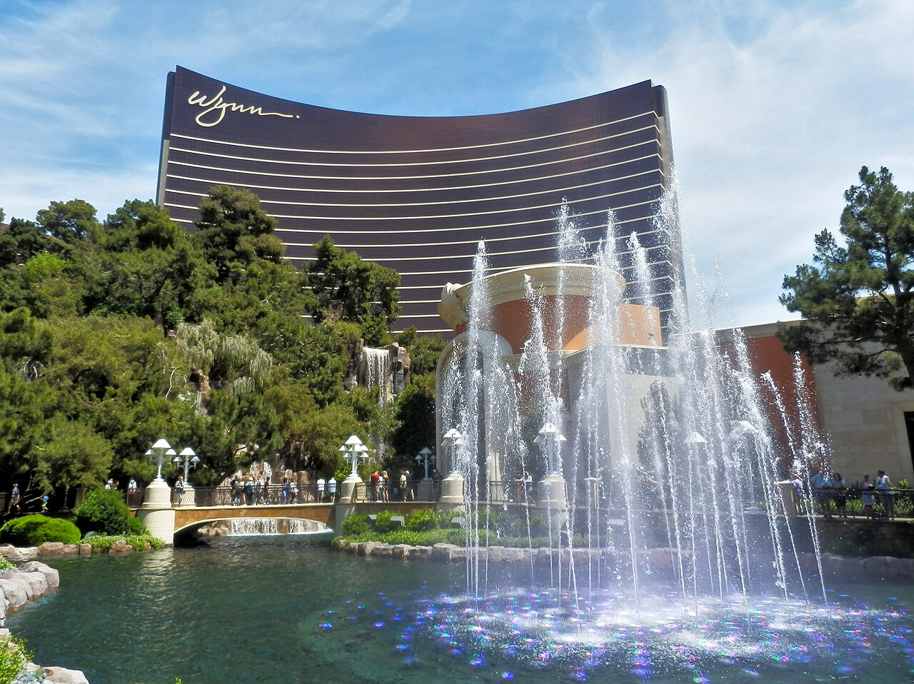 A curved bronze-glass hotel tower rising behind trees and a pool with tall fountain jets.