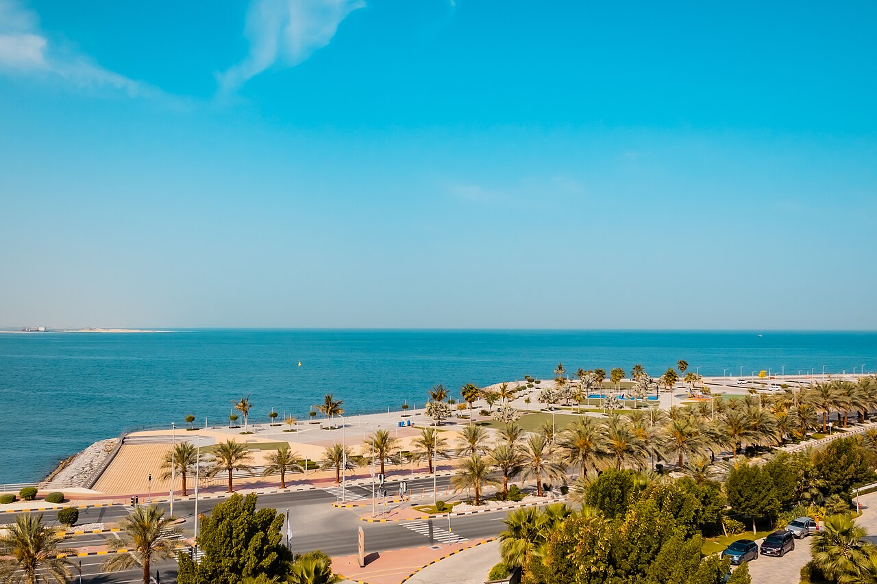 A palm-lined seafront promenade and beach beside bright turquoise water under a clear sky.