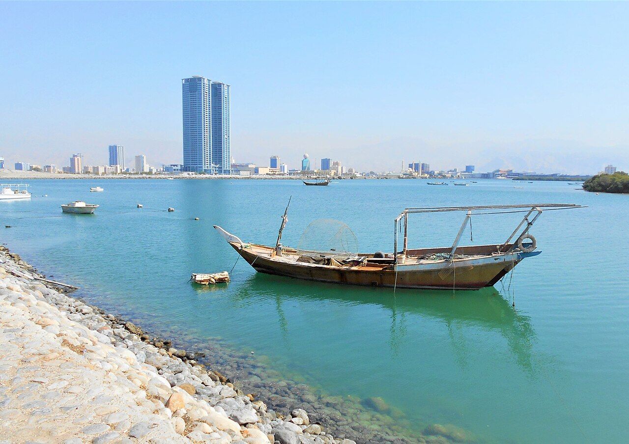 A wooden dhow at anchor in calm green water, with a rocky shore in front and towers on the far bank.